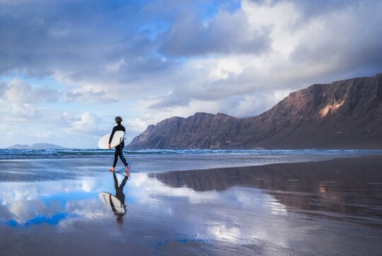 Un surfista con la tavola sulla sabbia bagnata di Playa de Famara, con la bassa marea